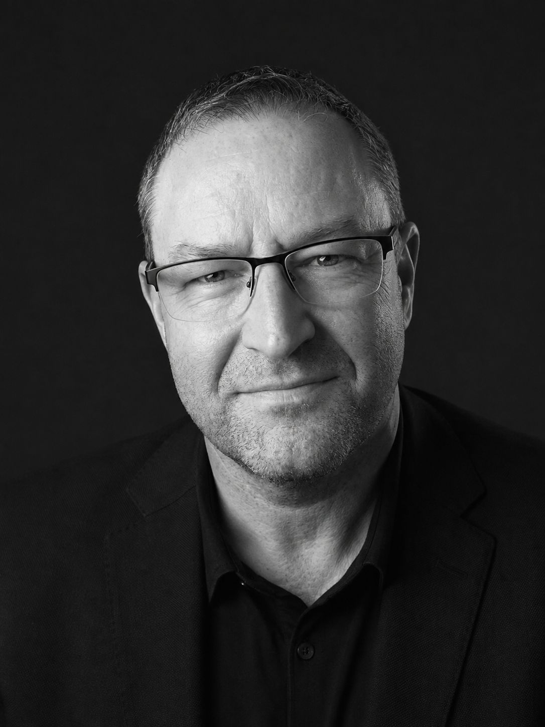 Forrest Andrew Jones in a black and white studio portrait, wearing a dark jacket and dark shirt, with half rim glasses and short hair.